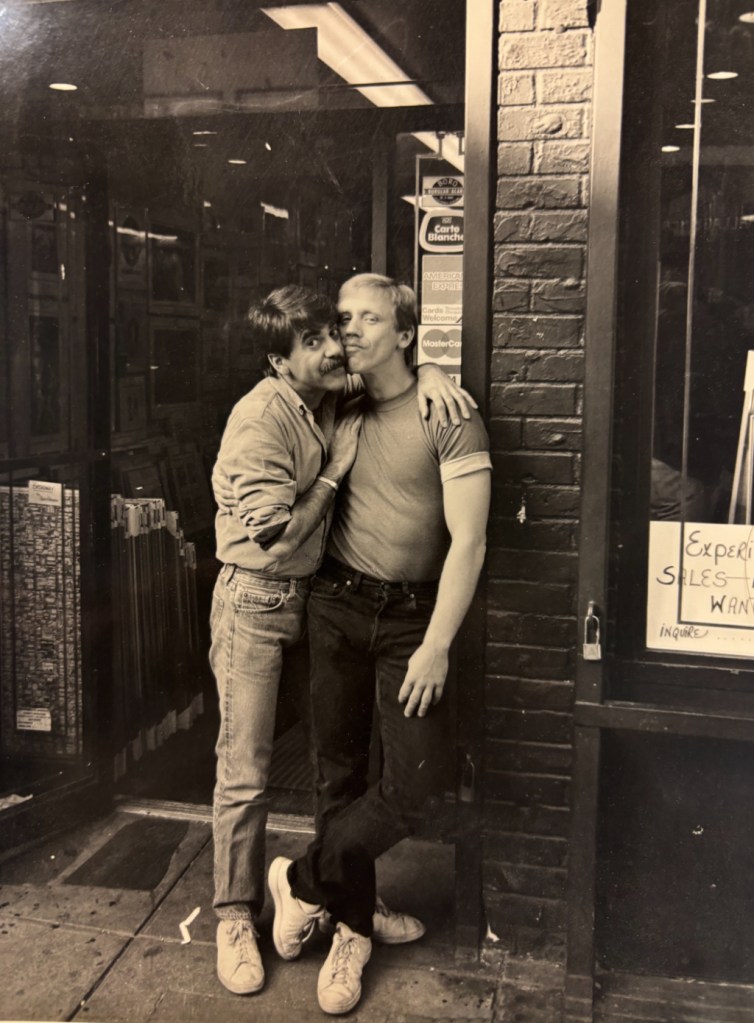 Black-and-white photo of Bill Hendricks outside All State Art on Christopher Street, NYC (1981/82), with a friend leaning in to kiss his cheek.